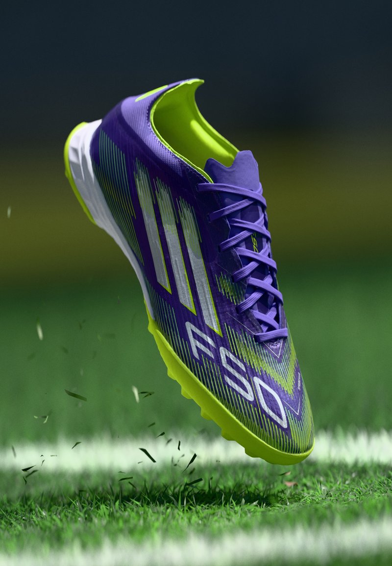 Scarpini da calcio viola e verde lime con una superficie testurizzata, logo prominente e design a tre strisce, sospesi sopra l'erba.