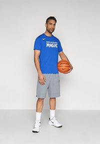 Blaues T-Shirt mit kurzen Ärmeln und dem Schriftzug "ORLANDO MAGIC", graue Shorts, hält einen orangefarbenen Basketball, trägt weiße Nike-Sneaker und Socken.