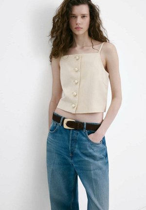 Joven con cabello rizado que lleva una camiseta beige sin mangas con botones y vaqueros azules de talle alto con un cinturón negro, de pie con las manos en los bolsillos.