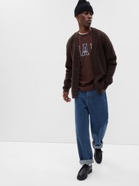 Brun cardigan over en mørkebrun sweatshirt med hvite detaljer, paret med oppbrettede blå jeans og svarte sko, toppet med en svart lue.