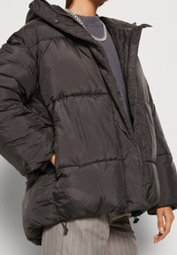 Chaqueta tipo abrigo en gris oscuro con un diseño acolchado, cierre frontal con cremallera, dobladillo ajustable con cordón y capucha. Tejido texturizado con acabado brillante.