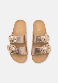 Metalliska roseguld slide-sandaler med två breda remmar, som har nubbade detaljer och stora cirkulära spännen på en beige fotsäng.