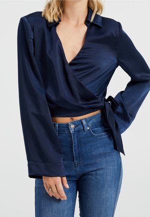 Vrouw draagt een navyblauwe satijnen wikkelblouse gestrikt aan de zijkant, gecombineerd met een high-waisted blauwe spijkerbroek.