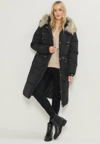 Abrigo acolchado negro con capucha forrada de piel sintética, de largo completo, diseño acolchado, cierre con cremallera y dos bolsillos frontales. Lleva puesto sobre un suéter ligero.