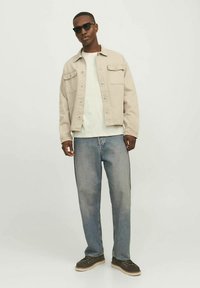 Béžová džínová bunda přes světle zelené tričko, doplněná volnými modrými džínami a tmavými botami. Doplňky zahrnují černé sluneční brýle.