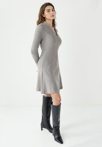 Grijze lange mouwen jurk met een getailleerde bovenkant, een uitlopende rok en een Mandarin-kraag, gecombineerd met hoge zwarte leren laarzen en stilettopumps.