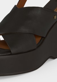 Μαύρο δερμάτινο πλατφόρμας πέδιλο με φαρδιές διασταυρωμένες λουρίδες και λουράκι στον αστράγαλο με κούμπωμα.