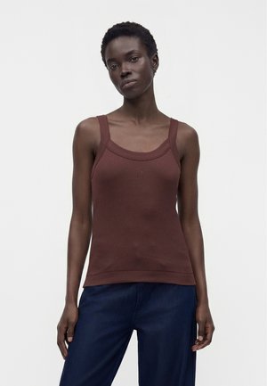 Personne mince aux cheveux courts et bouclés, portant un débardeur marron à encolure carrée sans manches et un pantalon bleu foncé, debout devant un fond uni.