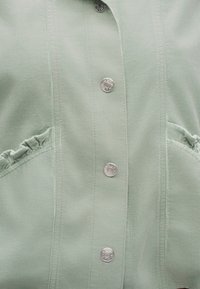 Υφασμα κορδούρας σε ανοιχτό πράσινο με κατακόρυφες ραφές, ασημένια κουμπώματα με πατιλέτα και βολάν κατά μήκος των κυρτών άκρων της τσέπης.