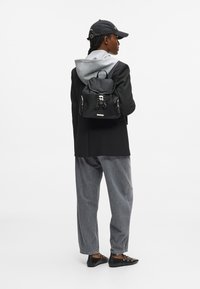 Άτομο που φοράει μαύρο καπέλο, μαύρο σακάκι, γκρι φούτερ, γκρι παντελόνι, μαύρα παπούτσια, και κρατάει μια μαύρη σακίδιο που κοιτάζει πλάγια σε λευκό φόντο.