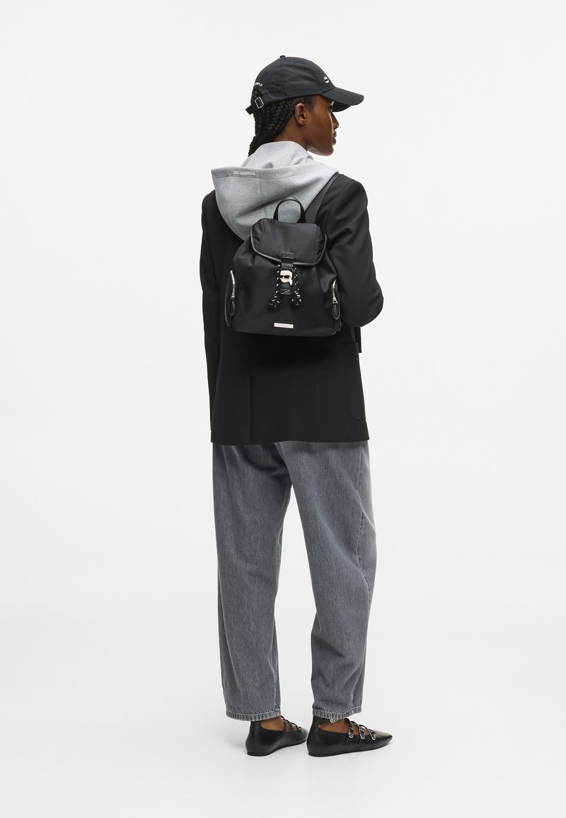 Άτο�μο που φοράει μαύρο καπέλο, μαύρο σακάκι, γκρι φούτερ, γκρι παντελόνι, μαύρα παπούτσια, και κρατάει μια μαύρη σακίδιο που κοιτάζει πλάγια σε λευκό φόντο.