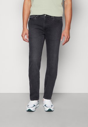 Mężczyzna w czarnych obcisłych dżinsach i białych trampkach stoi na jednolitym białym tle.