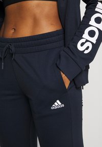 Gros plan d'une personne portant un pantalon de survêtement et une veste Adidas bleu marine, main dans la poche, avec le ventre visible et une partie du logo sur la manche de la veste.
