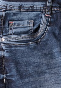 Tissu en denim bleu foncé avec une poche avant dotée d'un rivet en métal argenté, de coutures contrastantes et d'une finition lisse au toucher.