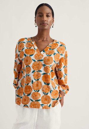 Žena s tmavými vlasmi viazanými v nízkom konskom chvoste, oblečená v bielej blúzke s oranžovým kvetinovým vzorom a voľných bielych nohaviciach na jednoliatom pozadí.