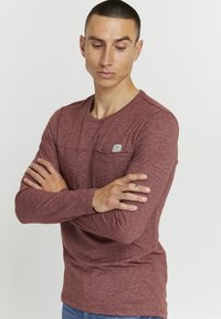 Langermet maroon t-skjorte laget av teksturert stoff, har en lomme på forsiden av brystet, rund halslinning, og en liten merkeetikett på lommen.