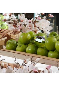 Une caisse en bois remplie de pommes vertes, une pomme métallique brillante, et des fleurs de magnolia artificielles roses et blanches autour de la caisse.