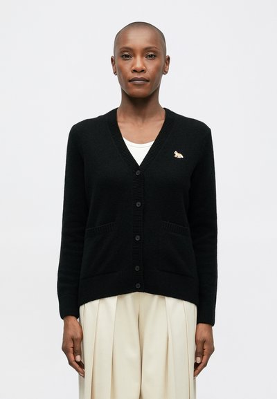 Cardigan noir avec un col en V, des boutons sur le devant et deux poches. Il présente une petite épingle dorée en forme de renard sur le coin supérieur gauche.