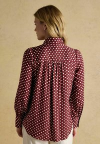 Red polka dot shirt with white dots, long sleeves, a collar, and gathered detailing at the back. The fabric appears smooth and lightweight.