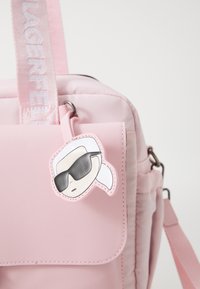 Ružičasti ruksak s sjajnim završetkom, s poklopcem džepa i privezakom u obliku crtanog junaka. Naglašen izdržljivim remenom s natpisom "LAGERFELD."