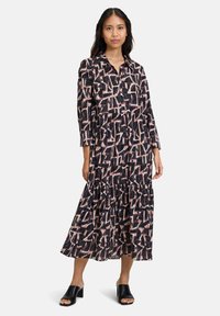 Schwarzes Kleid mit geometrischem Druck in Rosa und Creme, mit einem Knopfrückenkragen, langen Ärmeln und einem gestuften Rock. Tragen mit schwarzen Mules.