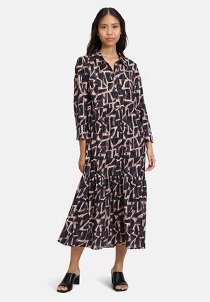 Schwarzes Kleid mit geometrischem Druck in Rosa und Creme, mit einem Knopfrückenkragen, langen Ärmeln und einem gestuften Rock. Tragen mit schwarzen Mules.