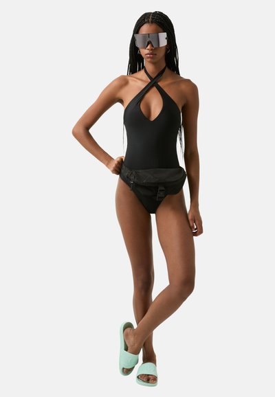 Black one-piece swimsuit with a deep V-neck and crossed straps; paired with a black waist bag and mint green slide sandals. Large sunglasses included.
