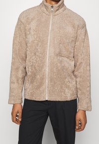 Beige fleecejakke med lynlås foran, høj krave og en brystlomme. Blødt, tekstureret stof med en afslappet pasform og lange ærmer.