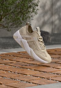 Chaussure de sport beige avec une tige en tissu texturé, une semelle coussinée et un logo embossé, présentant un design élégant et moderne avec un profil bas.