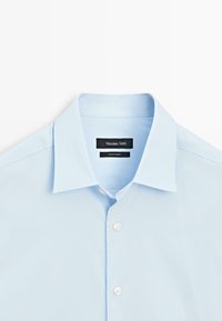 Lyseblå skjorte med klassisk krave, der har en glat tekstur, hvide knapper og et mærke med teksten "Massimo Dutti" indeni kraven.