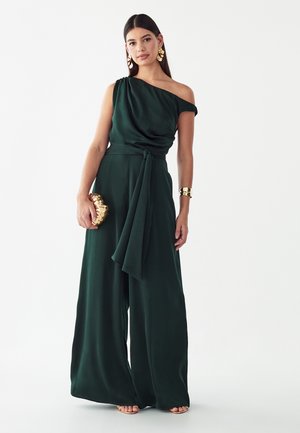Vrouw draagt een donkergroene jumpsuit met één schouder en wijde pijpen, houdt een gouden clutch vast en draagt gouden oorbellen en armband.