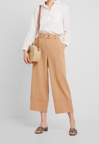 Beige weite Hosen mit hoher Taille und gefaltetem Saum, kombiniert mit einem weißen Hemd mit Knöpfen und einer geflochtenen Umhängetasche.