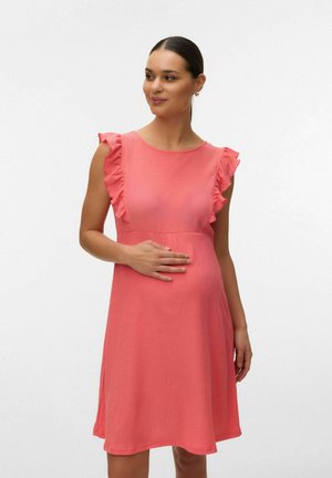 Kobieta w ciąży w koralowej sukience bez rękawów z falbanami na ramionach, ręka spoczywa na brzuchu, spogląda lekko w bok na jednolitym tle.