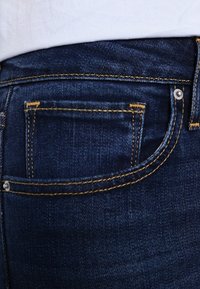 Jeans de mezclilla azul oscuro con costuras amarillas, un bolsillo trasero y un detalle de botón plateado. La tela tiene un acabado suave y ligeramente texturizado.