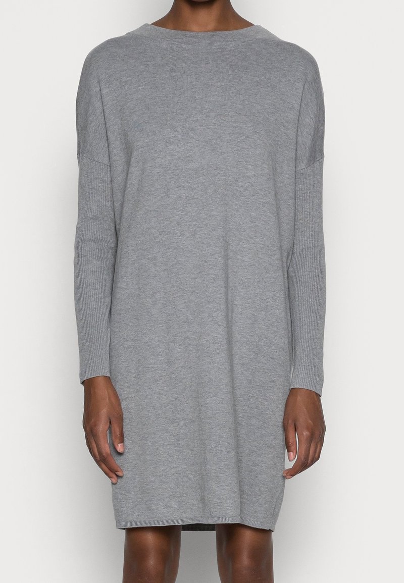 Personne portant une robe en maille grise, manches longues, longueur genou, avec un col rond, debout devant un fond uni.
