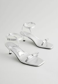 Silver metallic heeled sandals with a square toe, adjustable ankle strap, and a small decorative bow on the front. Block heel design.