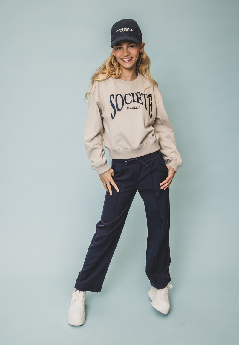 Lichtbeige sweatshirt met "SOCIETE" logo, marineblauwe tapered broek en witte sneakers. Draag met een marineblauwe baseballcap. Neutrale achtergrond.