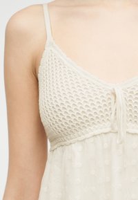 Κοντινό πλάνο ενός ατόμου που φοράει λευκό αμάνικο τοπ με πλεκτό πάνω τμήμα και διάφανο, πουα κάτω ύφασμα, δεμένο με μικρό φιόγκο στο κέντρο μπροστά.