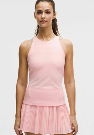 Kobieta z włosami związanymi do tyłu, ubrana w bezrękawnik w blady różowy kolor i pasującą plisowaną spódnicę, stojąca na tle jasnego, jednolitego tła.