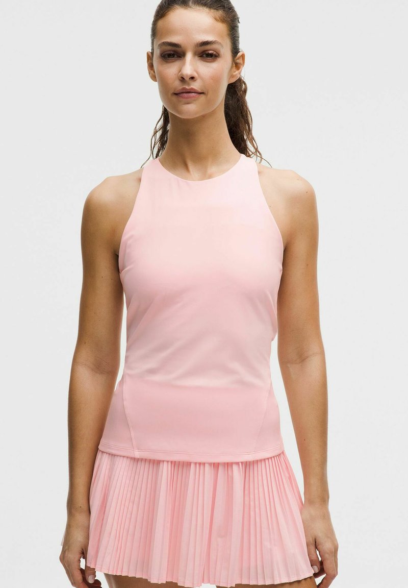 Mujer con el cabello recogido, vistiendo una blusa sin mangas de color rosa pálido y una falda plisada a juego, de pie frente a un fondo claro y liso.