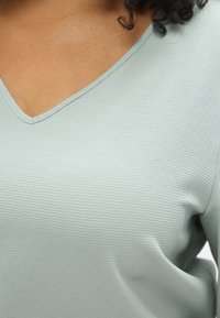 Ανοιχτό πράσινο τοπ σε σχήμα V με υφή υφάσματος, που διαθέτει διακριτικό κυματιστό σχέδιο και ομαλή επιφάνεια.