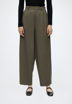 Femme portant un pantalon large taille haute vert olive, un haut noir à manches longues et des chaussures Mary Jane noires, debout sur un fond blanc.