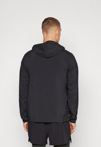 Sudadera negra con capucha, de mangas largas, textura suave y dobladillo trasero alargado. Lleva puesta sobre pantalones cortos oscuros, con un diseño minimalista.