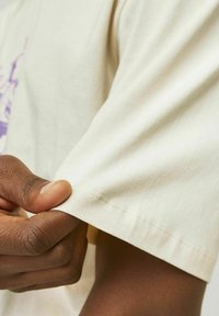Cream-colored cotton t-shirt with a smooth texture, featuring a short sleeve and visible stitching detail on the cuff.