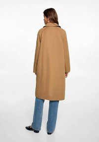 Cappotto in misto lana color sabbia con colletto a scollo, maniche lunghe e taglio dritto. Abbinato a jeans a gamba larga blu chiaro e scarpe nere.
