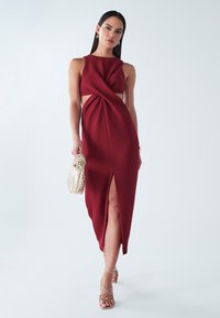 Bordowa sukienka bez rękawów z wycięciami, skręconym przodem i wysokim rozcięciem. Noszona z złotymi sandałkami na paskach i plecioną złotą torebką.