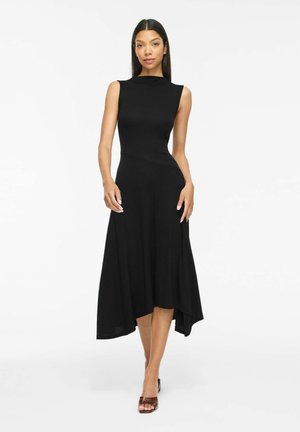 Femme aux longs cheveux foncés portant une robe midi noire sans manches avec un ourlet asymétrique, marchant vers l'avant en talons ouverts marron sur fond blanc.