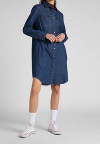 Jeanshemdkleid in dunkelblau, mit langen Ärmeln, durchgehender Knopfleiste, zwei Brusttaschen und geschwunghem Saum. Getragen mit weißen Sneakers und Socken.