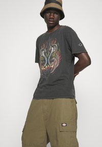 Camiseta gráfica negra con un diseño colorido de serpiente y alas. Usada con pantalones cortos de carga oliva y un sombrero tipo bucket bicolor.