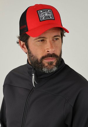 Homme à la barbe portant une casquette Oxbow rouge et noire et une veste noire zippée, regardant légèrement vers la droite sur un fond uni.